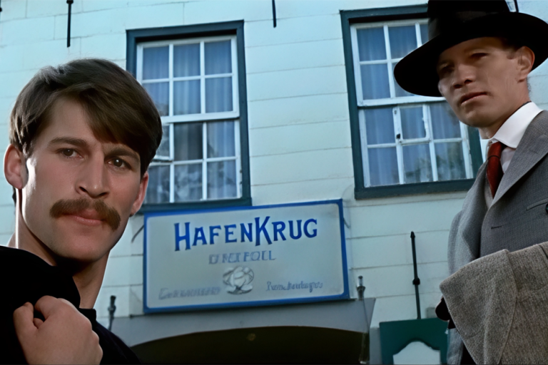 Standbild aus dem Film "The Riddle of the Sands" mit Simon MacCorkindale und Michael York. Zwei Männer in Anzügen und Hüten stehen vor einem Gebäude mit einem Schild 'HAFENKRUG BENSERSIEL'
