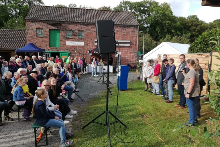 Gefeierte Aufführung der Krimikomödie "Joke & Harm unter Strom" des Oldersumer Theaterklottje am 14.9.2025 beim Museum Alte Seilerei in Oldersum. Mehrere Personen stehen nebeneinander auf einer Wiese vor einem Publikum, das auf Bänken vor einem Backsteingebäude sitzt.